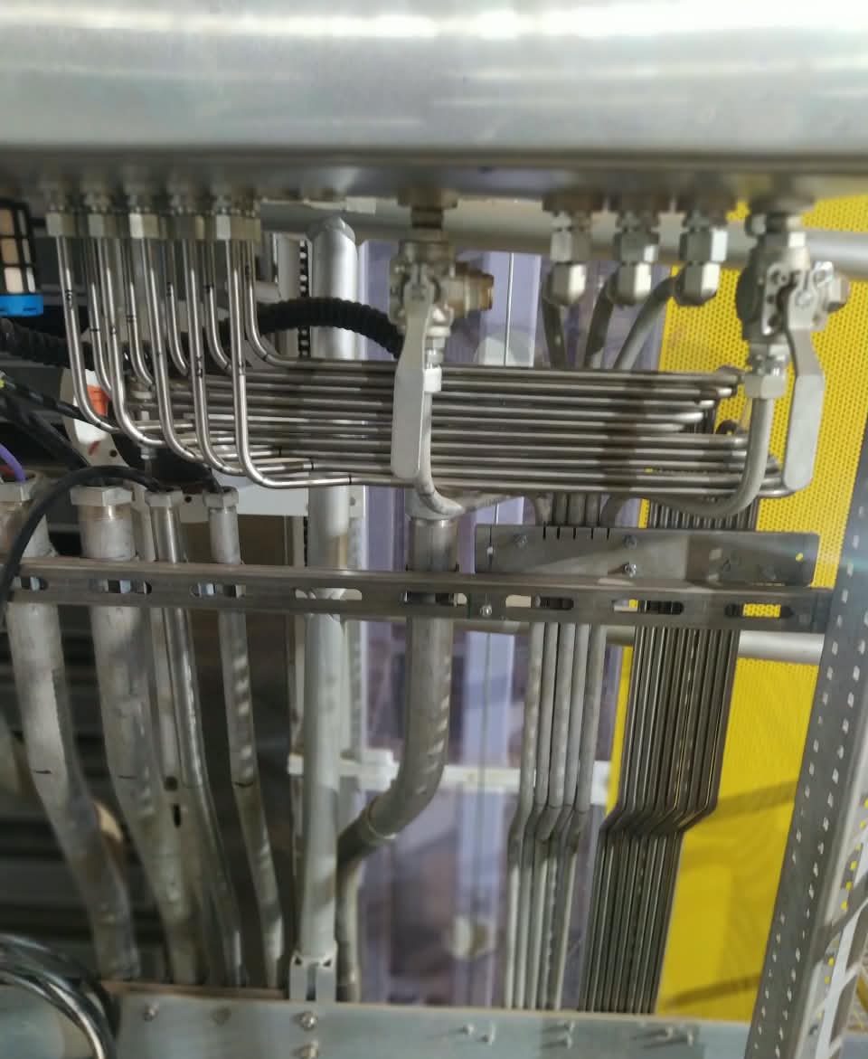 Fileira de tubos finos de aço inox presa por uma braçadeira, com uma estrutura amarela ao fundo
