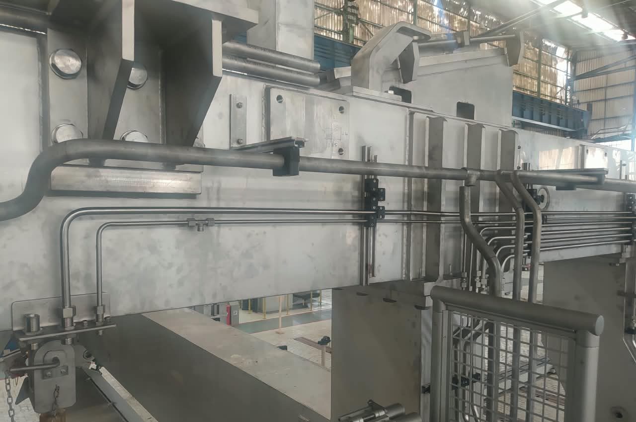 Tubos de aço inox acompanhando uma viga metálica, com o interior da fábrica ao fundo